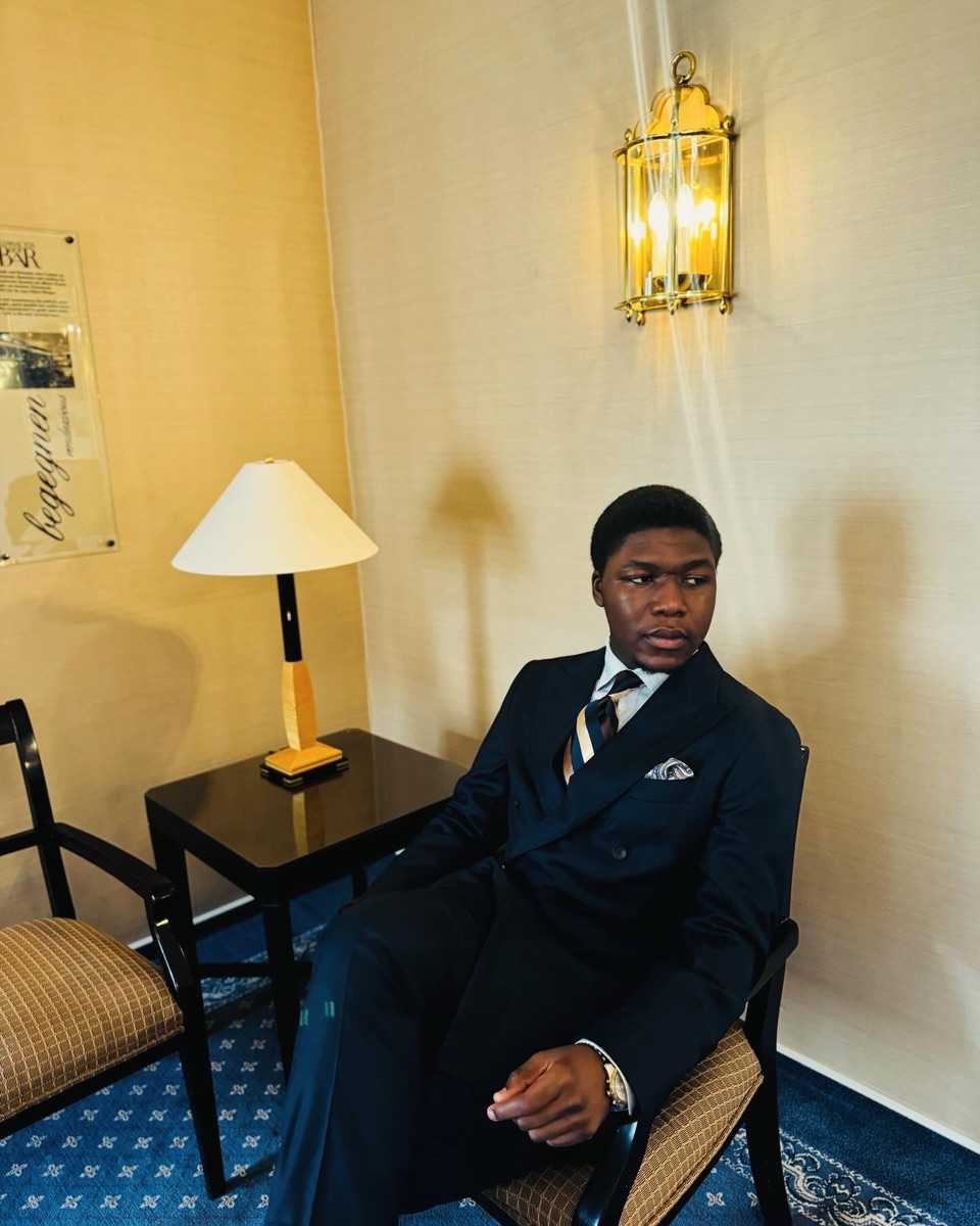 David seated in a double-breasted navy suit beneath a brass wall lantern